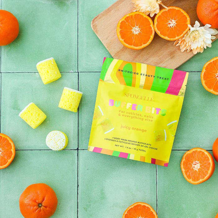 Spongellé Juicy Orange PediPOP Buffer Bits surrounded by fresh orange slices and yellow sponges on mint green tiles