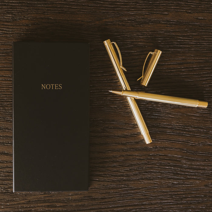 Black NOTES booklet with gold pens arranged on dark wood surface, styled desktop setting
